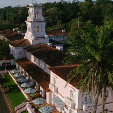 Aerial view of Hotel das Cataratas surrounded by lush greenery and palm trees, featuring a prominent white bell tower and multiple outdoor umbrellas along the terrace.