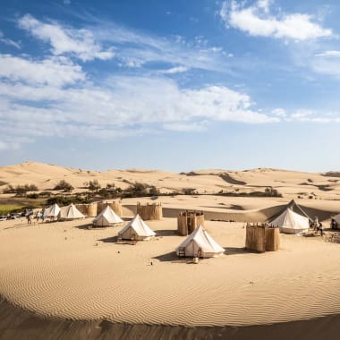 A desert dune camp with several white canvas tents arranged in a semi-circle surrounded by sand dunes under a partly cloudy blue sky.