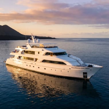 Eine weiße Luxusyacht namens 'Galapagos Explorer' fährt bei Sonnenuntergang auf ruhigem Ozeanwasser nahe vulkanischer Inseln.