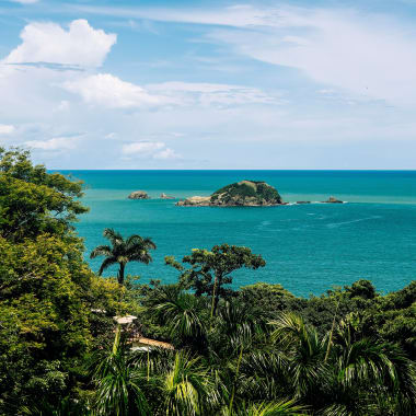 Lush green tropical forest overlooking a turquoise ocean with a small rocky island in the distance under a partly cloudy sky.