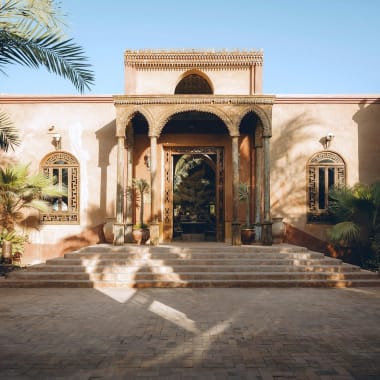 Großer Eingang zum Al Moudira Hotel mit traditioneller Architektur, Palmen und Steinstufen, die zur Tür führen.