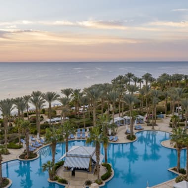 Eine luxuriöse Resort-Poolanlage mit zahlreichen Palmen, Sonnenliegen und einem Pavillon, Blick auf den Strand und das Meer bei Sonnenuntergang.