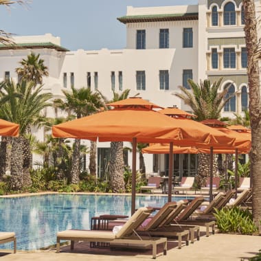 Outdoor swimming pool at Four Seasons Hotel Rabat with rows of lounge chairs shaded by orange umbrellas and palm trees, set against a white hotel building under clear sky.