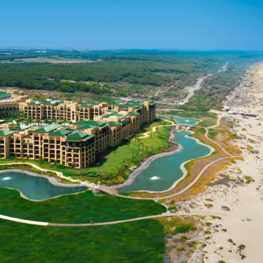 Ein großes Resort mit grünen Dächern, umgeben von angelegten Seen, grünen Feldern und Wegen, direkt neben einem langen Sandstrand mit Meereswellen unter klarem blauem Himmel.