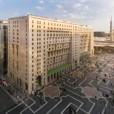 Außenansicht des Sofitel Shahd Al Madinah Hotels bei Tageslicht, mit einem großen Platz und Menschen, die sich bewegen.