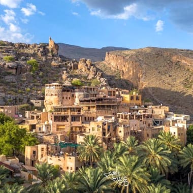 A hillside village with traditional mud-brick buildings, surrounded by lush green palm trees, set against a mountainous landscape under a partly cloudy sky.