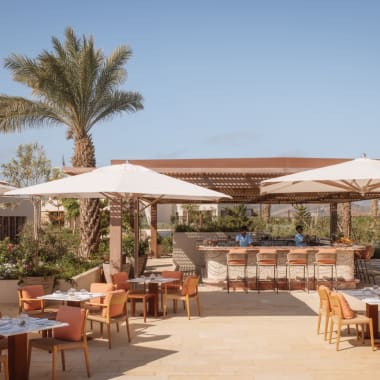 Outdoor-Restaurant mit Tischen, Stühlen, großen Sonnenschirmen, Barbereich und Poolblick bei sonnigem Wetter.