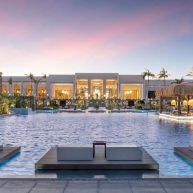 Luxushotel mit einem großen Swimmingpool, Liegestühlen und einem Gebäude mit Glastüren hinter Palmen bei Sonnenuntergang.