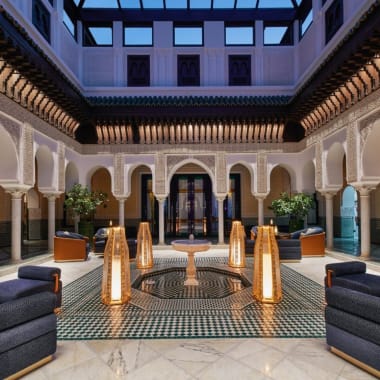 Elegant indoor courtyard with arched doorways, patterned floor tiles, and modern seating arrangements, illuminated by tall floor lamps.