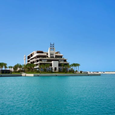 Ein modernes Hotel und ein markantes segelförmiges Gebäude am Wasser, mit klarem blauen Himmel und Wasser im Vordergrund.