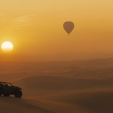 Ein Fahrzeug fährt über Sanddünen, während ein Heißluftballon am Himmel während des Sonnenuntergangs schwebt.