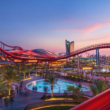 A vibrant amusement park during sunset with a large roller coaster, palm trees, and a swimming pool.
