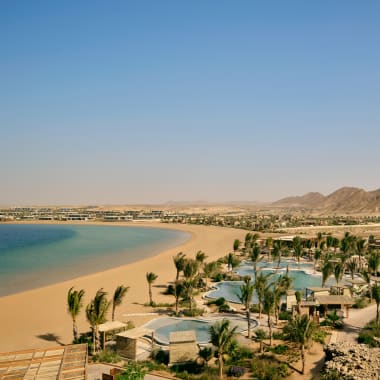 Curved sandy beach along a calm blue sea with a luxury resort featuring multiple pools and palm trees in a desert coastal setting, with mountains in the background.