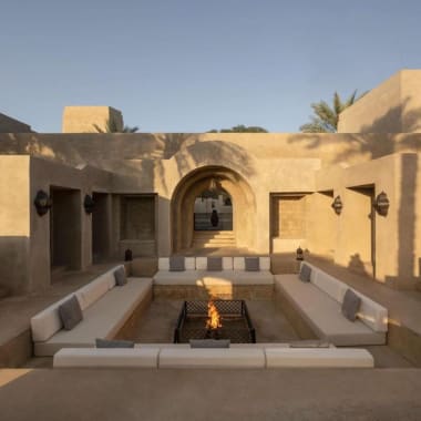 Ein luxuriöser Außenbereich mit einer Feuerstelle in der Mitte, umgeben von bequemen Sitzgelegenheiten, in einer Wüsten-Architektur mit sandfarbenen Mauern und Bögen.