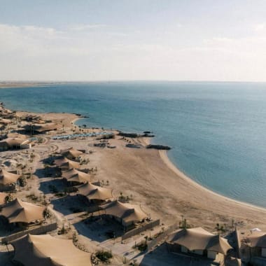 Luftaufnahme eines Strands mit beigen Zelten entlang des Sandufers, neben ruhigem blauem Wasser unter einem bewölkten Himmel.