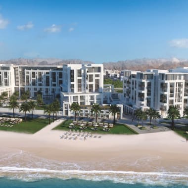 A modern beachfront resort with white buildings, surrounded by palm trees, on a sandy beach under a blue sky.