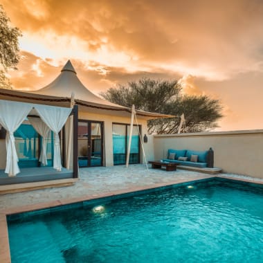 A luxurious poolside villa at sunset with a swimming pool, outdoor seating, and a tented canopy.