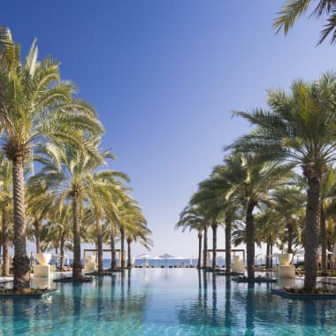 A luxurious infinity pool at Al Bustan Palace, a Ritz Carlton Hotel, surrounded by palm trees with a view of the sea in the background.