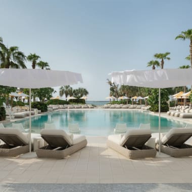 Luxuriöse Außenpool-Anlage in einem tropischen Resort mit weißen Liegen und Sonnenschirmen, umgeben von Palmen und klarem Himmel.