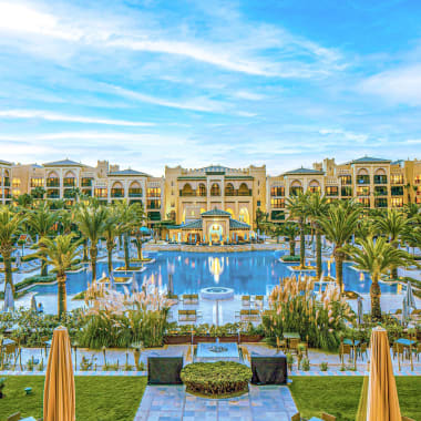 Luxurious resort with a large symmetrical outdoor pool surrounded by palm trees and multi-storey beige buildings under a partly cloudy sky.