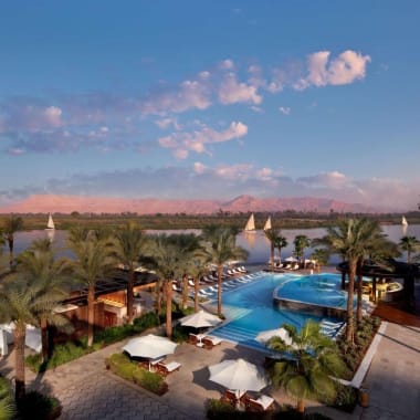 Luftaufnahme des Poolbereichs im Hilton Luxor Resort & Spa mit Palmen und Sonnenliegen, Blick auf einen Fluss und entfernte Berge.