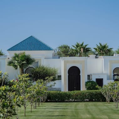 Resortgebäude mit blauen pyramidenförmigen Dächern hinter einem üppigen Garten mit Palmen und Orangenbäumen unter klarem blauem Himmel.