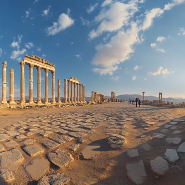 Ancient ruins with tall columns and cobblestone ground under a partly cloudy sky, with a few visitors in the distance.