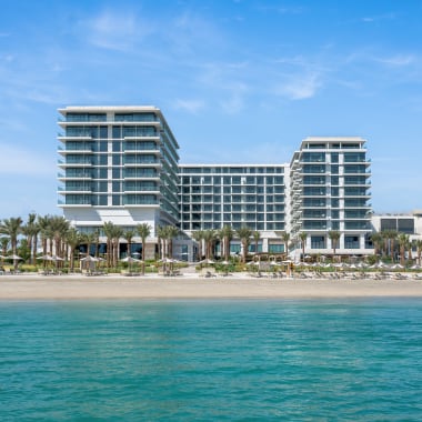 Ein modernes Strandhotel mit mehreren Stockwerken und Glasfassade, gelegen an einem tropischen Strand mit Palmen.
