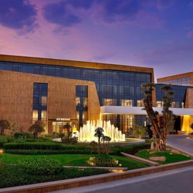 Außenansicht des Hotels St. Regis Riyadh mit beleuchteter Fontäne und gestalteten Gärten bei Dämmerung.