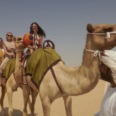 Eine Familie reitet auf Kamelen in einer Wüste, vorne steht ein Mann in traditioneller osmanischer Kleidung.