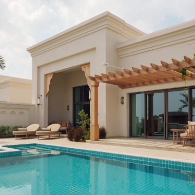 Moderne Villa mit privatem Swimmingpool, Sonnenliegen und Holzpergola, umgeben von Palmen unter bewölktem Himmel.