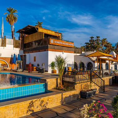 Außenansicht des Sonesta Nouba Hotels in Assuan mit Swimmingpool, Palmen und sonnigem Wetter.