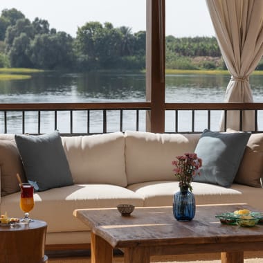 Gemütliche Loungemöbel im Freien mit einem weißen Sofa, grauen Kissen, Holztischen und einer Vase mit Blumen, mit Blick auf den Nil und grünen Hintergrund.
