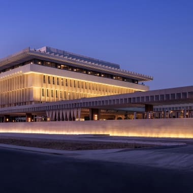 Das The Ned Doha Gebäude bei Dämmerung beleuchtet mit moderner, horizontaler Architektur und warmem gelben Licht.