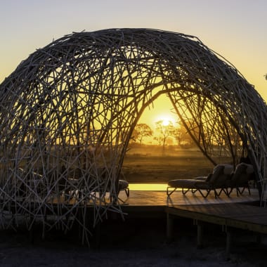 Eine kunstvolle, kuppelförmige Struktur aus ineinander verschlungenen Ästen, die bei Sonnenaufgang eine ruhige Lounge-Bereich mit Liegestühlen umschließt.