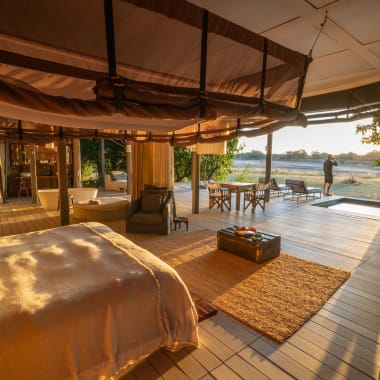 Luxuriöser Safari-Zelt-Innenraum mit großem Bett, Sitzecke und Blick nach draußen auf einen Sonnenuntergang über der Landschaft.