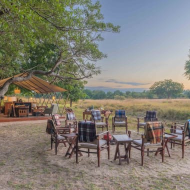 A campfire setup with chairs arranged in a circle in a natural field setting during sunset, with a tent and trees nearby.
