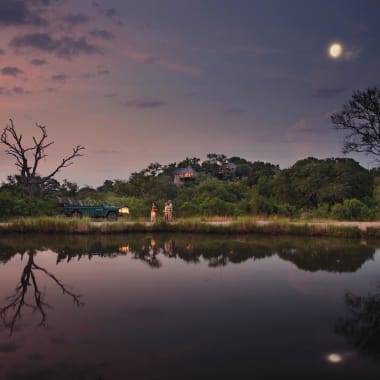 Eine ruhige Seenlandschaft bei Dämmerung mit Bäumen, Hügeln und dem Mond, der sich im Wasser spiegelt.