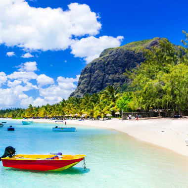 Ein lebhafter tropischer Strand auf Mauritius mit klarem türkisfarbenem Wasser, einem hellgelben und roten Boot nahe dem Ufer, mehreren Segelbooten am weißen Sandstrand und einem üppigen grünen Berg im Hintergrund unter einem teilweise bewölkten blauen Himmel.