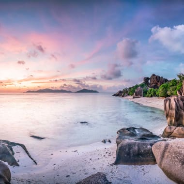 Ein unberührter Strand mit großen Felsen und üppigem Grün auf La Digue, Seychellen, bei Sonnenuntergang.