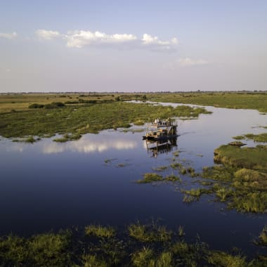 A boat navigates through a wide, calm river amid lush green wetlands under a partly cloudy sky.