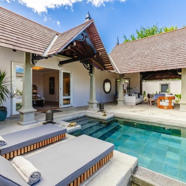 Luxusvilla mit privatem Pool, ausgestattet mit zwei grauen gepolsterten Liegestühlen und einem überdachten Terrassenbereich mit Pflanzen und Gartenmöbeln unter einem Ziegeldach.