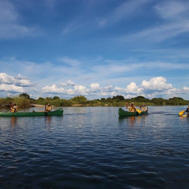 Vier Personen paddeln in einem ruhigen Fluss mit einem blauen Himmel und Wolken darüber.
