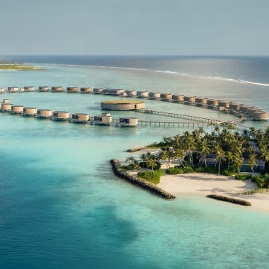 Luftaufnahme von luxuriösen Überwasser-Bungalows, die sich im türkisfarbenen Lagunenwasser der Malediven kreisförmig um eine zentrale Struktur gruppieren, mit weißem Sandstrand und Palmen im Vordergrund.