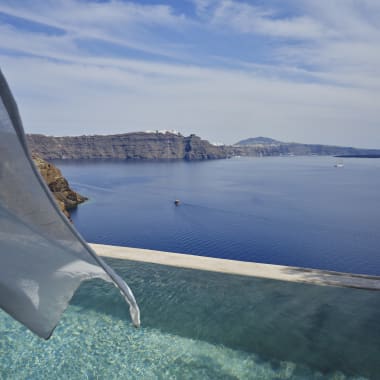Blick auf ein ruhiges blaues Meer mit felsigen Klippen und einem kleinen Boot, gesehen von einem Pool mit einem im Wind wehenden Vorhang.