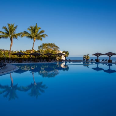 Ein großer Außenpool, umgeben von Palmen und Sonnenschirmen, mit Liegestühlen rund um den Pool an einem klaren, sonnigen Tag.