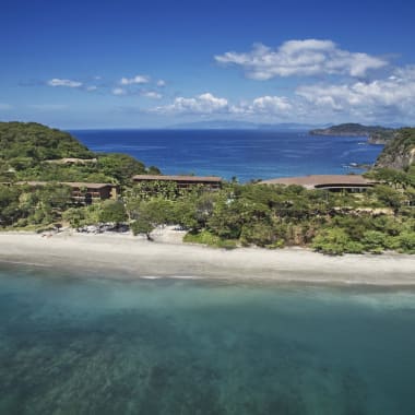 Aerial view of a coastal landscape with a beach, lush greenery, and buildings set among trees under a partly cloudy sky.