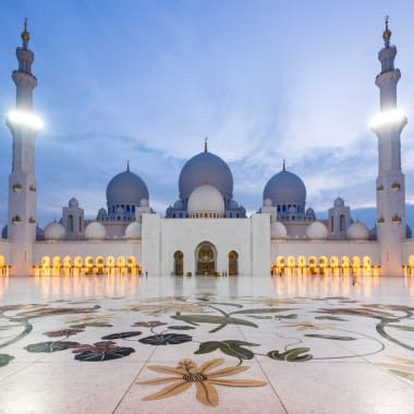 Ein majestätischer Blick auf die Sheikh Zayed-Großmoschee mit weißen Kuppeln und Minaretten bei Sonnenuntergang, reflektiert im Marmorhof mit Blumenmosaik.