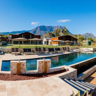A modern outdoor swimming pool area with multiple lounge chairs and umbrellas, set against a backdrop of wooden cabins and a mountainous landscape under a clear blue sky.
