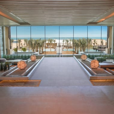 Luxuriöse Hotellobby mit großen Glasfenstern, die auf einen Strand mit Palmen blicken.